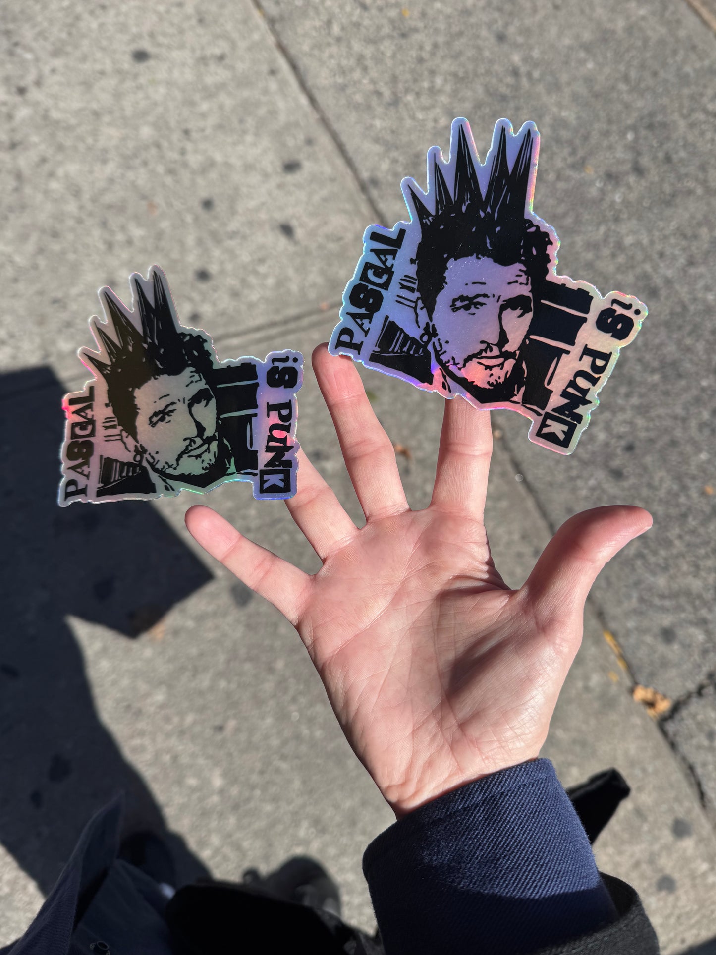 Hand holding two stickers with a stylized portrait of Pedro Pascal with a punk hairstyle and a pavement background
