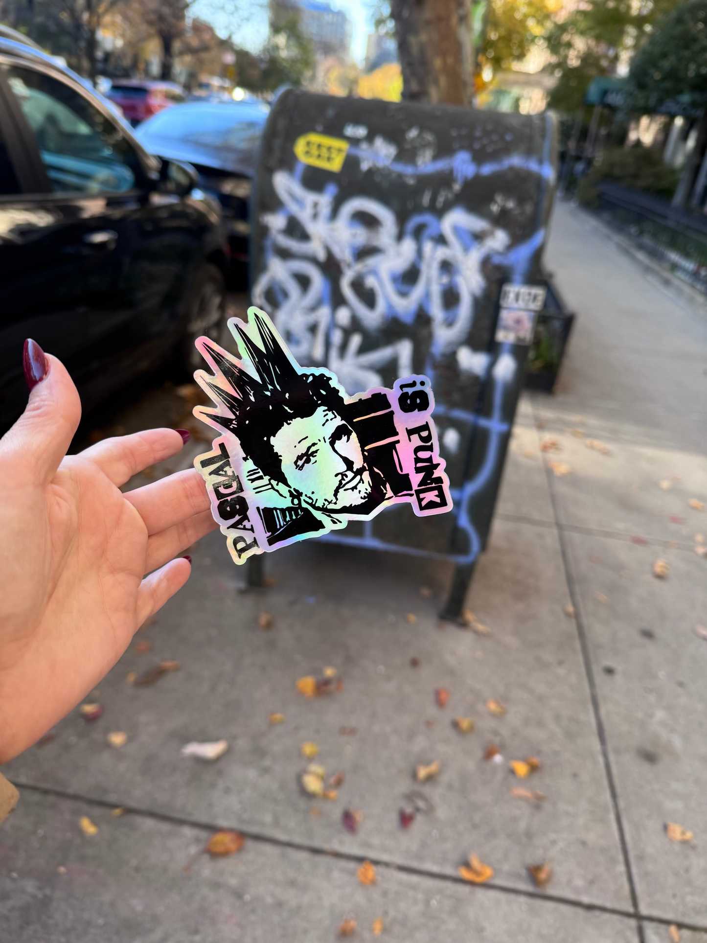 Hand holding a colorful sticker with a depiction of Pedro Pascal with a punk hairstyle in front of a mail dropbox with graffiti.