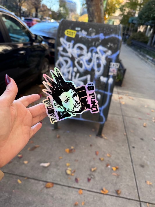 Hand holding a colorful sticker with a depiction of Pedro Pascal with a punk hairstyle in front of a mail dropbox with graffiti.